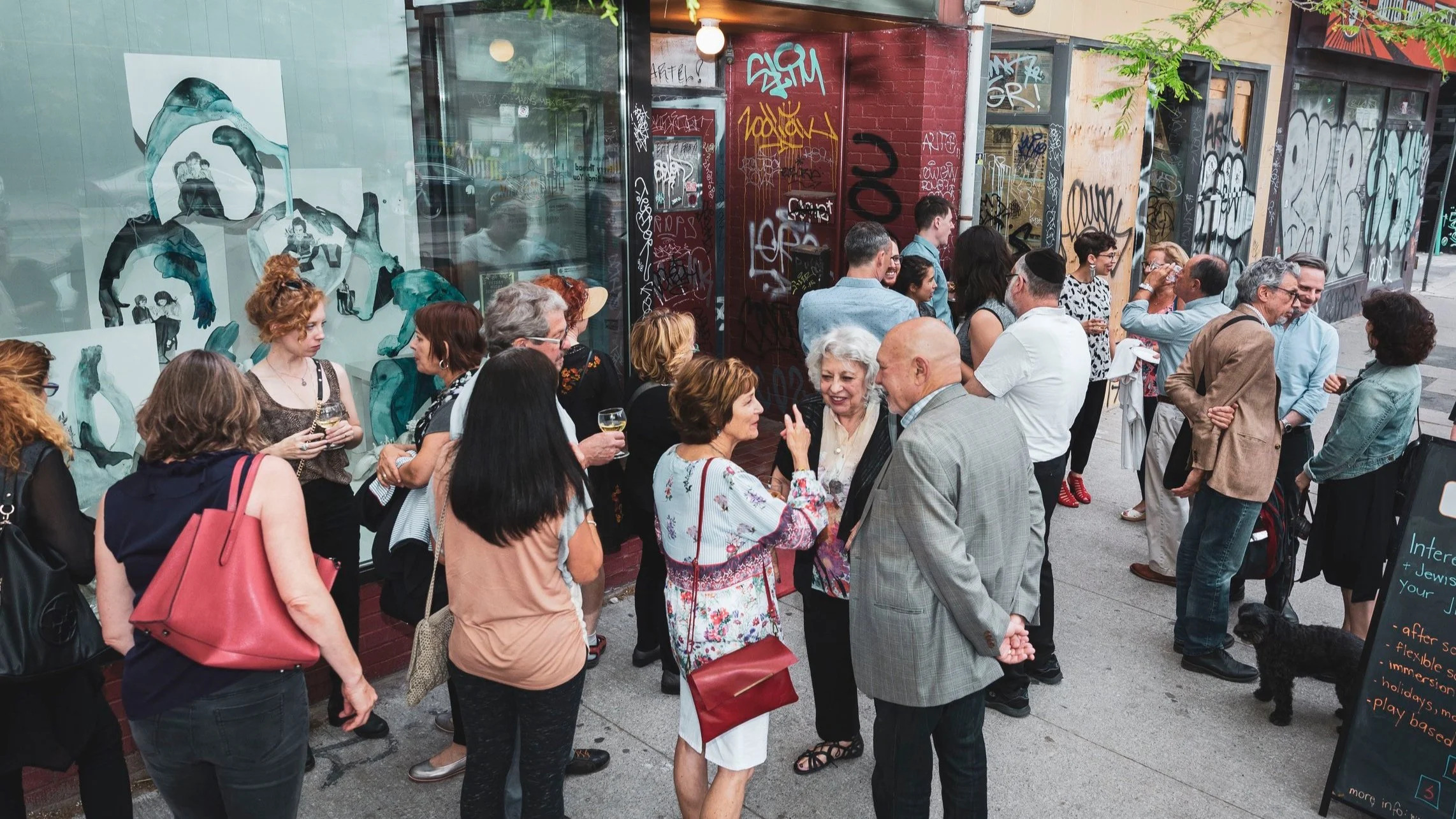 gallery opening with dozens of individuals standing on a sidewalk outside in front of a storefront with graffiti on its walls.